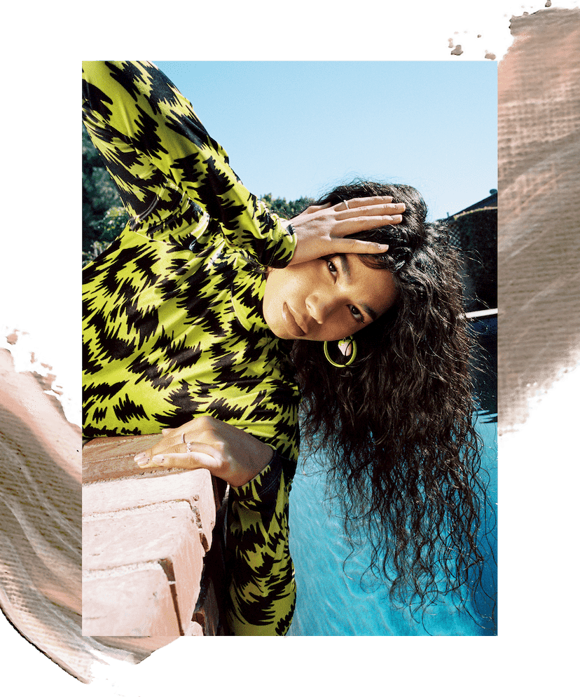 Model Ariel Toole is wearing a neon yellow and black printed blouse and holding her head over the pool. She has long wavy brown hair. In the second image, the same model is wearing black-and-white polka dotted shorts, black heeled sandals, and a black and neon yellow top. She is swinging her hair to one side. There is an illustration of brown and peach paint swatches behind the images.