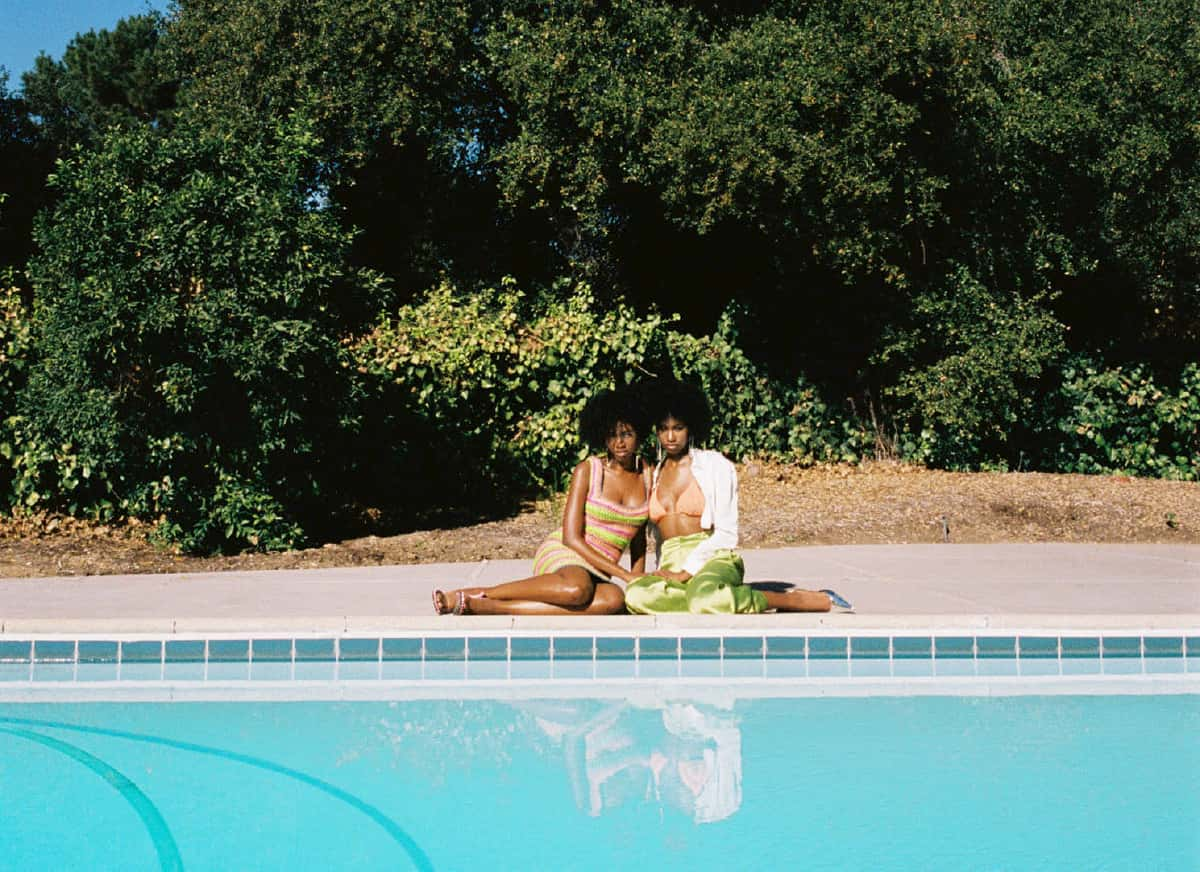 Twins Alana and Amaya January sit near the edge of the pool with a large tree behind them. One is wearing a green skirt, pink bikini top, and white button down. The other is wearing a crochet dress.